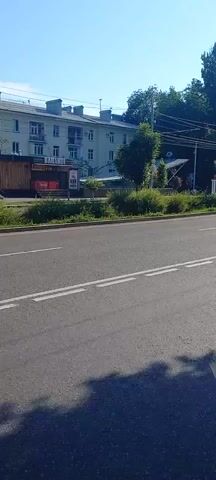 The video shows a road from the perspective of someone standing on the sidewalk. The road is paved and has white lane markings. There is a sidewalk next to the road, made of gray pavers. On the sidewalk, there is a parking sign indicating parking is allowed between 8:00 and 19:00. A dark-colored car is parked on the side of the road, just before the parking zone. There are trees and bushes on the side of the road opposite the sidewalk. In the background, there are buildings and more trees. The sky is blue and clear, suggesting it is daytime. The video pans slightly to the right, showing more of the road and the parked car.