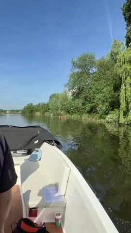 From a boat, people are visible standing in shallow water near the riverbank. The boat is moving, and the environment consists of a river, trees, and greenery along the riverbank. The sky is clear and blue.