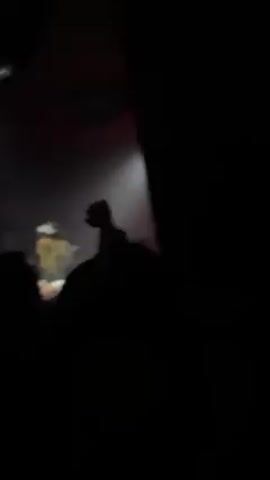 The video shows a dimly lit nightclub environment with a DJ performing. Initially, the view is obscured by people in the foreground, but it soon clears to reveal the DJ with long blond hair and a black hat, who is standing behind a DJ console with another individual next to them. The background features large black speakers framed with purple and white. A spotlight illuminates the DJ, and the crowd is visible in silhouette, with at least one person raising a hand in the air. The atmosphere appears lively, with music playing.