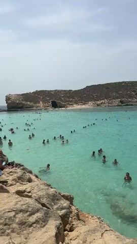 The video captures a lively scene at a sun-drenched beach, likely located in Għajnsielem, Malta. The turquoise waters are filled with a variety of people, suggesting a popular tourist destination during warm weather. The time of day appears to be mid-day, with clear skies and bright sunshine casting a warm glow over the landscape.

The primary subjects are the numerous individuals enjoying the water. They are engaged in swimming, wading, and playing, with some seen floating on inflatable rings. The diverse group of people includes adults and children, all seemingly relaxed and having a good time. Among the objects visible are several boats anchored in the distance, indicating boat tours or private vessels present in the bay. The natural setting is characterized by rocky shorelines and a barren, hilly landscape in the background, typical of Mediterranean coastal regions.

The activity is clearly recreational, centered around swimming and enjoying the beach environment. The clear, vibrant blue of the water is a striking feature, suggesting a clean and inviting swimming area. The overall mood conveyed is one of fun, relaxation, and summer enjoyment. There are no visible text elements within the video.