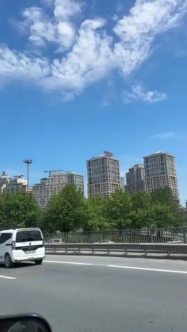 The video shows a view from a moving vehicle on a highway on a sunny day with scattered clouds. Buildings are visible in the distance behind a line of green trees. One building is labeled "Venezia Outlet". There are street signs and guardrails along the highway, and other vehicles pass by. The sound of wind is prominent in the audio.