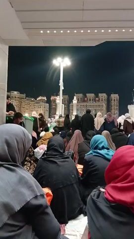 Here's a description of the video:

The video shows a large group of people, mostly women, sitting on the ground in what appears to be an outdoor space near a mosque. The women are wearing various colors of hijabs and abayas.

In the background, there are tall buildings and white minarets illuminated at night. There are lampposts providing additional light to the area. 
The people seem to be observing or listening to something that is happening outside the frame, as suggested by the audio.