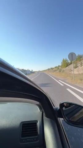 This is a first-person perspective video of someone driving a car on a highway. The camera is pointed forward, showing the road ahead and the hood of the car. The sky is clear and blue, and the landscape on either side of the road is dry and hilly, with sparse vegetation. There are guardrails and fences along the side of the road. The car is traveling on a paved road with white lane markings. The timecode indicates the video is approximately 5 seconds long, showing a continuous forward motion.