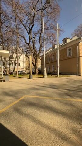 The camera shows a city square on a sunny day, with bare trees and buildings as a backdrop. Several people are walking through the square. One man in a black jacket walks past a bus stop, talking on his phone. Later, a woman helps a young boy ride a red and blue scooter. There are a few parked cars nearby, and streetlights are visible. Yellow painted lines mark the pavement in the square. The overall scene is quiet and typical of an urban park area.