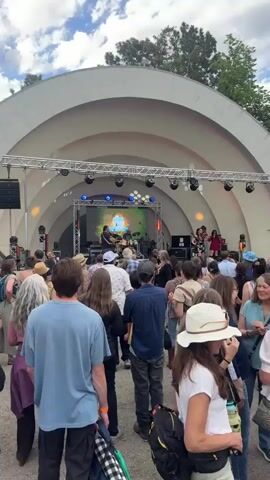 A crowd of people is watching a band perform on a stage under a large arched canopy. The band includes guitarists, a drummer, and two women playing violins. Stage lights are visible above the performers. The crowd consists of individuals of varying ages and appearances, with most facing the stage. The environment appears to be an outdoor concert or festival setting.