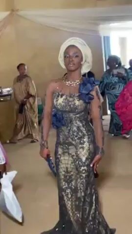 The video shows a young woman in a long black and gold lace gown, with a white headwrap, standing in a crowded indoor space. The gown has an asymmetrical neckline and blue floral embellishments. She is carrying a small blue clutch. Other people are visible in the background, wearing a variety of traditional African attire. The setting appears to be a celebration or event, possibly a wedding. The room has a neutral color scheme, with beige walls and white drapery hanging from the ceiling. There is buffet-style food service set up in the background. The camera is handheld and moves slightly as the videographer focuses on the woman.