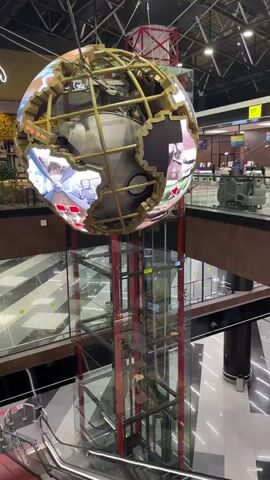 The camera faces down into the lower levels of an open-air building. Two people walk on an escalator that descends to the level below. Above the escalator hangs a large digital globe of sorts, showing images on its surface. Above the lower level where the escalator ends, there is a clear glass elevator with red supports. The flooring on the lower levels is black and white checkered. In the background, some people are seated in chairs in a waiting area.