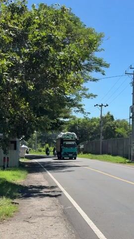 The camera is stationary, showing a two-lane road in what appears to be a tropical environment. The sky is clear and blue. On the left side of the road is a large green tree. To the left of the tree is a small structure with a tent pitched in front of it. The road itself has a yellow stripe down the middle. A turquoise truck drives down the right lane of the road towards the camera, loaded high with what appear to be bags of something. Three motorcycles follow the truck, passing in front of the camera. The background includes power lines and a concrete wall.