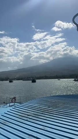 The video pans from a blue and white striped deck, showing a life preserver attached to a railing. In the distance, a wide expanse of water reflects the bright sun and scattered white clouds in a blue sky. Several dark-colored boats, possibly ferries, are visible on the water. Beyond the water, a lush green landscape slopes upwards towards a large, misty mountain. The scene conveys a peaceful, sunny day at sea with a prominent mountain backdrop.