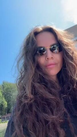 A woman with long, curly brown hair and sunglasses takes a selfie outdoors. She is wearing a black top. The background shows a bright blue sky, green trees, and a body of water with mountains in the distance. The video appears to be taken from a boat or similar vessel, as indicated by the movement of the water and the perspective.