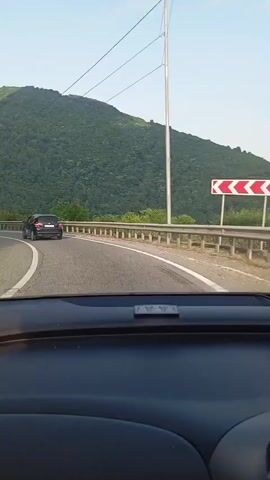 The video shows a view from inside a car driving on a paved road. The road curves to the left, with a metal guardrail along the right side. A dark-colored car is visible ahead, also driving on the road. On the right side of the road, a large, tree-covered mountain dominates the landscape. On the left side of the road, the terrain slopes upwards, also covered with trees and some rocky outcrops. Utility poles with wires run along the right side of the road. The sky is clear and blue, with the sun appearing to be lower in the sky, casting shadows. The interior of the car is visible in the foreground, including the dashboard.