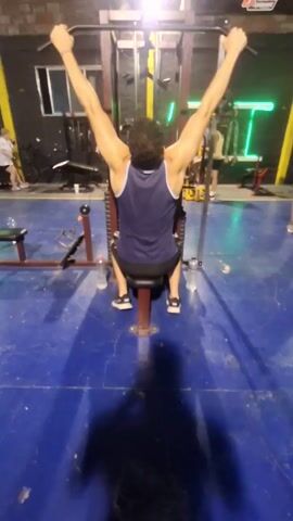 The video shows a person doing seated lat pulldowns in a gym. The person is wearing a blue tank top and dark shorts, sitting facing away from the camera on a padded seat attached to a lat pulldown machine. Their hands grip a horizontal bar suspended above them. The machine has a metal frame and the weights are obscured. There are two water bottles on the floor next to the person. The gym has a blue floor and is lit by overhead lights. In the background, other gym equipment and people are visible. The camera is positioned behind the person performing the exercise, capturing their back and arm movements. The shadow of the person filming is visible in the foreground.