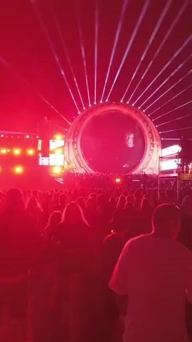 A large, circular screen displaying a DJ performing at a concert. The screen is illuminated with red lights, and there is a crowd of people in front of it. The overall atmosphere is lively and energetic.