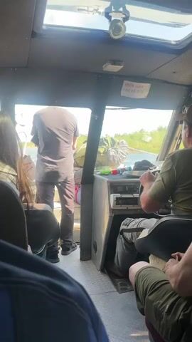 The video shows the interior of a boat with several passengers. In the foreground, blurry figures are seated. In the middle ground, a man is standing and looking out the front window. Another man is seated at the helm of the boat, steering. Outside the window, there is water and vegetation. There is a piece of paper with writing on it attached to the ceiling of the boat.