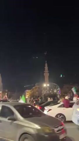 At night, a street scene is captured in a low-angle video, depicting cars moving through a crowded street. Many people are seen on the roofs of the cars. They are carrying flags, some of which appear to be white with black lettering, and others are green. The crowd appears celebratory, given the festive atmosphere. The street is lined with buildings illuminated by streetlights. A building with Arabic text is also visible. In the background, a mosque with a single minaret and a brightly lit, spherical structure can be seen. The sky is dark, suggesting it is late in the evening or night. The camera appears to be handheld, as there is slight movement throughout the video.