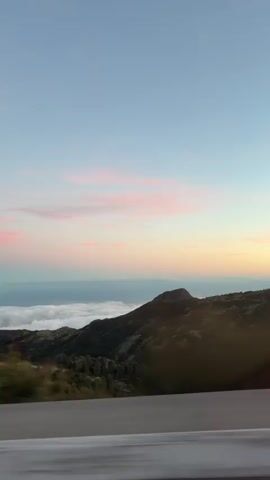 The video shows a landscape at sunset. The camera is moving, likely from a vehicle on a road next to a railing. There's a mountain range with vegetation in the foreground. Beyond the mountains, a sea of clouds is visible, topped by what appears to be the ocean. The sky is pale blue with streaks of pink and orange from the setting sun. Trees pass by the camera in a blur as the vehicle moves.