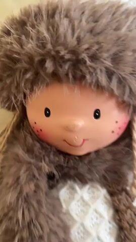 A close-up shot of a doll wearing a brown furry hat, a matching furry scarf, a white knitted top with a brown criss-cross pattern, and brown overalls with two wooden buttons. The doll has a round, pale face with rosy cheeks, freckles, and painted blue eyes and a smiling mouth. The doll's hair appears to be made of stringy brown material, and its lower body and legs are covered in white and brown knitted material, with the legs crossed. The background is a textured, light brown surface, possibly a couch or upholstery.
