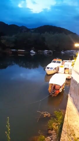 The video shows a body of water with several boats docked along a riverbank at dusk. The water is calm and reflects the sky and the silhouettes of mountains and trees on the opposite shore. The sky is a deep blue with some lighter patches near the horizon. The boats are of various sizes and designs, ranging from small motorboats to larger, multi-tiered vessels. Some of the boats are tied to the riverbank, while others are moored further out in the water. The riverbank is made of stone and concrete, and there are some plants and vegetation growing along the edge. There is no visible human activity. The lighting is dim, with only a few artificial lights visible on the boats and along the riverbank.