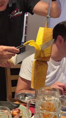 A server wearing a black shirt with a "Picanha" logo is carving a piece of grilled pineapple from a large skewer. The skewer holds two rectangular blocks of pineapple, one above the other. The server uses a knife to slice a piece from the bottom block of pineapple, and a pair of tongs is used to hold the skewer steady. A man in a white t-shirt is seated to the right, looking at the pineapple. In the foreground, there are plates with meat and glasses of beer. The scene appears to be a restaurant setting.