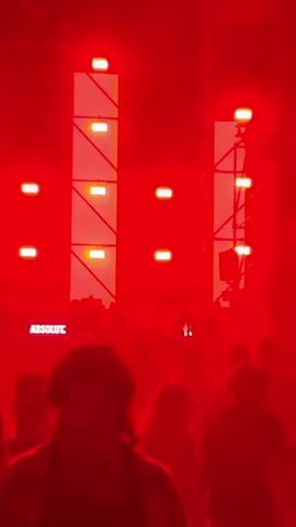 The video shows a nighttime outdoor event, likely a concert or festival, with a large crowd of people visible. The stage area is illuminated by bright, dynamic lighting, predominantly red in the first segment and transitioning to blue and white in subsequent segments. The lighting appears to be synchronized with the music, creating a pulsating effect. A DJ booth or performance area is positioned behind a table with a prominent "ABSOLUT" logo, suggesting a sponsorship. Several figures, presumably performers or DJs, are visible behind the table, interacting with equipment. The crowd is dense and appears to be actively engaged with the performance, with silhouettes of individuals dancing and looking towards the stage. Tall, scaffolding-like structures are present behind the stage, housing additional lighting and possibly audio equipment. The overall atmosphere is energetic and immersive, characteristic of a live music event.