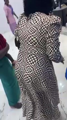 Here is a detailed description of the video:
A woman with dreadlocks and a checkered dress dances indoors with other women and a man.
The woman in the checkered dress is facing away from the camera at the start of the video, but turns to face the camera and points to it. She is smiling.
There are at least two other women, one wearing green pants and holding a small child in a red cloth, and another in a blue dress.
A man stands in the background filming with a phone. There are other people visible further in the background, and audio of people singing. 
The room is white with a glossy floor.