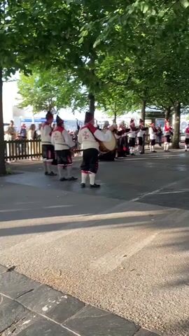 A group of musicians in traditional attire are performing outdoors. The subjects are people dressed in white shirts with red vests adorned with a cross symbol, black pants with red trim, and black hats. They are playing various instruments, including drums and what appear to be bagpipes. The scene is set in a park-like area with lush green trees providing shade. Sunlight casts long shadows on the paved ground, indicating it's likely late afternoon or early morning.

The activity is a musical performance, possibly a parade or a public celebration given the festive attire and outdoor setting. The musicians are arranged in a line, facing towards the right of the frame. Some individuals in the background, dressed in civilian clothes, appear to be watching the performance. The overall mood seems cheerful and lively, conveyed by the musical activity and the sunny weather.

A sign with the word "GIJÓN" is visible in the background, confirming the location as Gijón, Spain. The weather appears to be pleasant and sunny. The attire of the performers suggests a cultural event or a traditional folk performance.