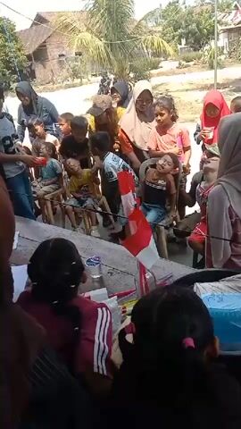 The video shows a group of children and adults outdoors, possibly at a community event or celebration. Many children are seated on chairs in a row, facing a central area. Some adults are standing behind the children, observing or interacting with them. One child in the foreground, a girl with dark hair, is visibly distressed or unwell, with her head tilted back and eyes closed. Another child near her also appears to be having difficulty. A small Indonesian flag, red and white, is visible near the children. The environment appears to be a rural or semi-rural area, with a building and vegetation in the background. Some children are holding what appear to be snacks or toys. The overall atmosphere seems to be one of a gathering, but with a moment of concern for the children who are not feeling well.