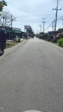 The video shows a first-person perspective of someone riding a motorcycle down a paved road lined with buildings and utility poles. The sky is cloudy, and the sun is shining. The road appears to be in a somewhat rural or suburban area, with low-rise buildings on either side. There are a few other motorcycles visible in the distance. The camera is positioned low, suggesting it is mounted on the handlebars or dashboard of the motorcycle. The scene is consistent with everyday traffic in a tropical or subtropical region, possibly Southeast Asia, given the palm trees and style of buildings.
