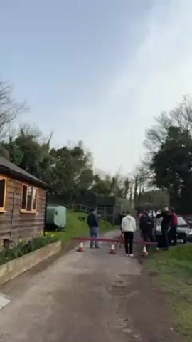 The video shows a group of people standing behind a barrier constructed of red and white cones, with a red bar connecting them. Behind them are several cars parked. The group is looking at a grassy knoll. A small, wooden structure is to the left. A countdown is heard from three to one. A loud bang is heard, and white smoke and sparks shoot up into the sky from the knoll.