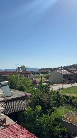 This video captures a sunny day with a clear blue sky and a bright sunbeam visible in the upper portion of the frame. The scene looks out over a residential area with several houses visible, many featuring terracotta tiled roofs. In the foreground, a rooftop with a solar water heater is prominent, along with corrugated roofing material. Lush green trees and bushes, some with vibrant purple bougainvillea flowers, are scattered throughout the landscape. In the mid-ground, more houses and a paved road are visible. Beyond the houses, a body of water, likely the sea or a large lake, stretches towards a distant, hazy treeline or mountain range. Several boats, including what appear to be fishing vessels, are moored on the water. The overall impression is of a peaceful, rural or coastal setting on a warm, clear day.