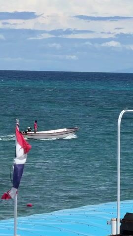 A small white boat with a red stripe along its side is moving across the water, powered by an outboard motor at the stern. A person wearing a pink shirt stands in the boat, facing towards the stern. The boat is leaving a wake behind it. In the foreground, a portion of a blue structure, possibly the deck of a larger boat or pier, is visible. To the left, a portion of the Thai flag, with its red, white, and blue stripes, is unfurled. The sea is a vibrant turquoise color, with visible waves and ripples. The sky above is a mix of blue and white clouds, with a distant landmass visible on the horizon. A bird is in flight near the water's surface.
