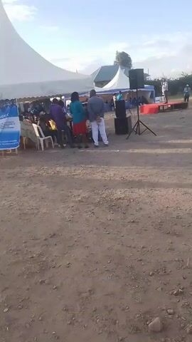 The video depicts an outdoor event under several large white tents, set on a dirt ground. People are scattered throughout the area, some seated in white chairs under the tents and others standing around. A small, raised platform covered in red carpet is visible, with a person standing on it addressing the crowd, supported by a speaker on a tripod. There is signage visible on one of the tents, and buildings are faintly visible in the background. The sky is partly cloudy, suggesting a fair weather day.