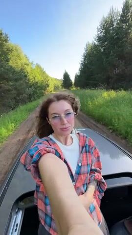 The video begins with a close-up selfie taken by a young woman with long, wavy brown hair, wearing glasses, a white t-shirt, and an open red and blue plaid shirt. Her arm is extended, holding the camera. She is leaning out of a car sunroof, with a wooded landscape visible in the background. A dirt road stretches ahead, flanked by green grass and tall trees. The woman smiles and turns her head to look out at the scenery as the car appears to be in motion. The sky is clear and blue, indicating daytime.