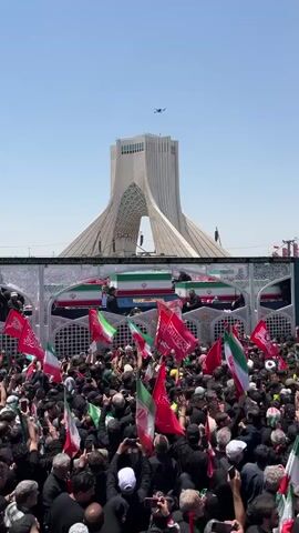 The video displays a crowd of people gathered outdoors, many holding flags. In the background, a distinctive modern structure resembling a tower is visible. In the foreground, there are several coffins draped with what appear to be national flags. People in the crowd are holding up their phones, seemingly to record or take pictures. A drone is visible in the sky above the tower. The atmosphere appears to be somber, indicated by the flags and the presence of coffins. The scene is illuminated by daylight.