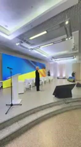 The video captures an indoor scene within a large, well-lit hall in Irpin, Ukraine, likely a conference or event space. The front of the room features a raised stage area with a white podium and a microphone stand. Behind the stage, a prominent large screen displays the distinct blue and yellow colors of the Ukrainian flag.

Initially, a single individual in dark clothing stands near the podium. As the camera pans to the right, it reveals a group of approximately twenty to thirty people, mostly men, standing in rows facing the stage. Many of these individuals have their right hands placed over their hearts, indicating a moment of reverence. The Ukrainian national anthem is playing in the background, strongly suggesting a formal ceremony or patriotic event.

The hall is brightly illuminated by recessed ceiling lights and decorative chandeliers. The flooring is a light-colored tile, and the ceiling has a grid pattern. The participants appear solemn and respectful. No discernible text is visible on the screen or other objects within the frame. The scene conveys a sense of national pride and formality.