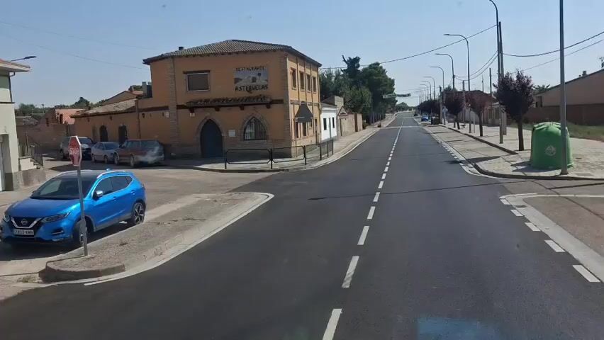 The video opens with a view of a two-lane road in what appears to be a small town. On the left side of the road is a two-story building with a sign that reads "RESTAURANTE ASTUELAS". There are several cars parked in front of the restaurant. The road is lined with trees and streetlights. A blue car is parked on the left side of the road, and a green dumpster is on the right. The camera pans to the right, showing more of the street and buildings. A banner hangs across the road that reads "FELICES FIESTAS!". The video continues to show the road stretching into the distance, with more houses and trees lining the sides. The scene is bright and sunny.