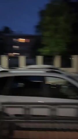 The camera, positioned behind a dark metal fence, captures a street scene at night. A white Hyundai taxi with a yellow stripe and a "Yandex Taxi" sign is parked on the side of the road, facing the camera. The car's headlights are on, and its license plate is visible. Streetlights illuminate the surrounding trees with bright green foliage. Another white car is partially visible behind the taxi. The sounds of an engine revving are prominent throughout the video.