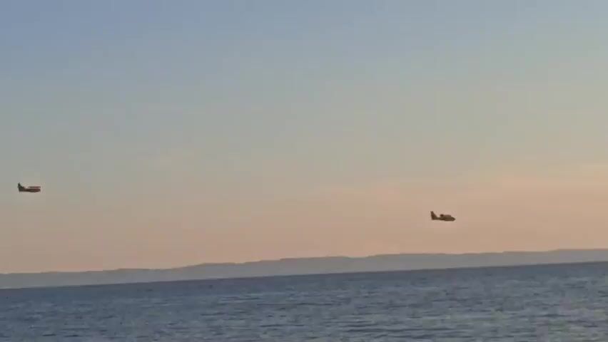 The video shows two amphibious aircraft flying over a body of water, likely the sea, near a landmass in the distance. The sky is clear with a gradient of colors from light blue at the top to a pale orange or pink near the horizon, suggesting it is either sunrise or sunset. The water is a deep blue with small ripples on the surface. The aircraft appear to be the same model, with their engines mounted above the wings and a distinctive high wing design. They are flying at a similar altitude and are spaced apart. One aircraft is positioned on the left side of the frame and the other on the right. The overall mood of the video is peaceful and serene, highlighting the vastness of the sky and sea. The context provided indicates the location is Sithonia Municipal Unit, Greece, which is a peninsula in northern Greece known for its scenic coastline and clear waters. The presence of amphibious aircraft might suggest a region where they are used for firefighting or search and rescue operations, especially given the warm climate of Greece and potential for wildfires. However, there is no visual evidence of any such activity in this specific clip.