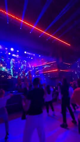 The video captures a concert or performance with a live audience dancing. The stage features a large screen displaying a cosmic or nebula-like visual. Red and blue stage lighting illuminates the scene, with lines of red lights running across the ceiling. People in the audience are visible from behind, with some raising their hands. The overall atmosphere appears energetic and festive.