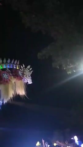 This video captures a lively night-time celebration in Dukuh Klopo, Indonesia. The scene is dominated by a large, elaborate float, likely part of a parade or festival. The float is adorned with colorful lights, intricate decorations, and what appear to be representations of mythical creatures or traditional figures, possibly inspired by Barong or similar cultural performances. Long, flowing tassels in shades of yellow and white hang from the float, adding to its dynamic appearance.

In the foreground, a crowd of people, many wearing matching blue shirts with text that is partially visible but appears to include "LG" and "LIMA," are gathered. The mood seems festive, with people observing the spectacle and some interacting with the moving float. The lighting is dynamic, with bright spotlights and colorful LEDs illuminating the scene against the dark night sky. Trees with lush foliage are visible, suggesting an outdoor setting, possibly a street or open area within a village. The time of day is clearly night, with the darkness punctuated by the vibrant lights of the celebration. No specific weather conditions are discernible, but the overall atmosphere is energetic and celebratory.