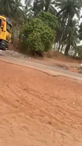 The video shows a yellow dump truck driving on a paved road alongside a dirt area. The truck has "MARIJAL" written on the side and a logo with a winged horse. The environment appears rural, with palm trees visible in the background. As the camera pans, we see people walking along the road near the truck. The dirt area is littered with debris, including tires and a damaged metal structure. The sky is overcast.