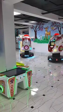 The video pans across an indoor amusement arcade. The floor is tiled with a white marble pattern and black squares. On the walls are colorful murals depicting cartoon characters and nature scenes. Several arcade machines are visible, including two futuristic-looking shooter games with lights, a foosball table, a claw machine, and a ride-on airplane for children. Two people are walking in the background, appearing to be customers. The overall environment suggests a family-friendly entertainment venue.