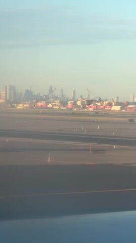 An aerial shot captures an airport runway and a distant cityscape under a clear sky. The runway is paved with asphalt and marked with yellow and white lines. Orange and white traffic cones are visible along the runway. In the background, a city skyline with numerous buildings and construction cranes can be seen. A large industrial area with red and white cranes and shipping containers is also in the distance. The footage appears to be taken from an airplane window.