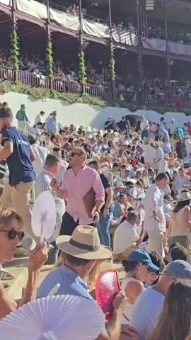 The video captures a bustling scene within an outdoor amphitheater, likely a bullring, filled with a large crowd of spectators. The people are seated on tiered bleachers under a bright, sunny sky. Many attendees are dressed in summer attire, with a noticeable number wearing hats and some holding fans, suggesting warm weather. The crowd appears diverse in age and gender. In the foreground, several individuals are visible, some walking and others seated. A man in a pink shirt and sunglasses is gesturing with his hand while holding a brown folder. Another man in a dark blue shirt is standing nearby. In the middle ground and background, rows of spectators fill the seating areas, many of whom are looking towards an unseen focal point, implying an event is taking place or about to begin. The architecture of the amphitheater is visible, with wooden railings and covered sections in the upper tiers. The overall atmosphere appears lively and engaged.
