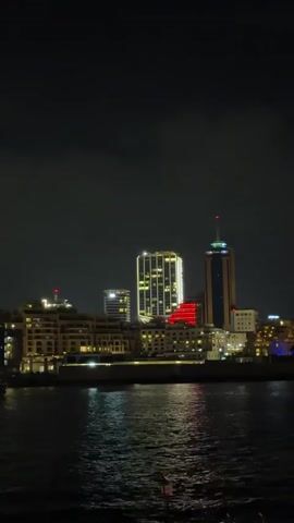 The video shows a nighttime view of a waterfront city skyline. Tall buildings with illuminated windows are visible, reflecting on the dark water in the foreground. The sky is dark, suggesting it is late at night or early morning. There are no people or discernible objects or actions in the frame.