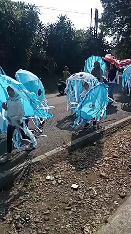 A group of people are walking down a road. The people are dressed in costumes that resemble jellyfish. The jellyfish costumes are light blue and white and have tentacles. Some of the people are carrying umbrellas that are also decorated with jellyfish designs. The road is paved and there is a curb on the right side of the road. There are trees and foliage on the left side of the road and in the background. The sky is visible and appears to be blue. The sun is shining. One person is inside a larger jellyfish costume that appears to be a mobile stroller. The other people are walking alongside the stroller. A motorcycle is parked on the left side of the road.