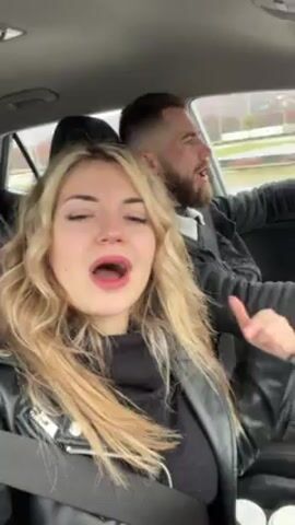 A young blonde woman in a black leather jacket lip-syncs to music in the passenger seat of a car. A bearded man is driving. They are both wearing seatbelts. The woman gestures to the camera a few times. The video is shot from inside the car. It is daytime.