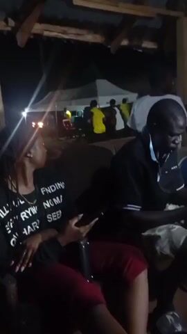 The video shows a low-angle, static shot from a covered outdoor area at night. In the foreground, a woman is seated on the left, wearing a black t-shirt with white text that reads "STILL I TRY EVERYTHING ON HARDER BUT EVERYTHING SEEMS TO BE FINDING ME AGAIN NO MATTER." She has dark hair pulled back and is looking to her left, holding a dark object in her hands. To her right, a man is seated, wearing a black polo shirt and patterned shorts. He is looking down and also appears to be holding something. Behind them, further in the background and slightly out of focus, are several other people standing under a white tent. Some are wearing yellow shirts. There are lights visible, suggesting it is an event or gathering. The environment appears to be rustic, with visible wooden beams and a corrugated roof overhead.