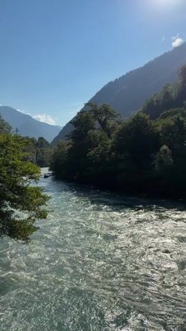 A fast-flowing river with white water rapids moves through a valley. The river is surrounded by dense green trees, with larger, darker mountains in the background. The sky is clear and blue, with the sun shining brightly in the upper right corner, casting a slight haze over the scene. The camera is positioned at an angle looking down at the river, with trees on the left side of the frame.