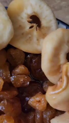 The video displays a close-up view of a plate filled with food items. The subjects are primarily food, including what appear to be fried dough pieces shaped like rings or fritters, and a generous portion of raisins. There are also darker, rolled pieces of what might be dried fruit leather or similar confection. The food is presented on a plate with a blue and white decorative rim. The setting appears to be an indoor environment, suggested by the lighting and the background texture which might be a countertop. The lighting seems to be artificial, with a warm tone. No people are visible. The activity taking place is simply the presentation of the food. There are no discernible location cues beyond the provided context of Baghramyan, Armenia. No text is visible in the video. The overall impression is a close-up, still life of a collection of sweet food items, likely ready to be served or consumed. The texture of the fried dough suggests it might be slightly greasy or glazed. The raisins vary in color from golden to a darker brown. The rolled dark pieces have a layered, almost wood-like appearance.